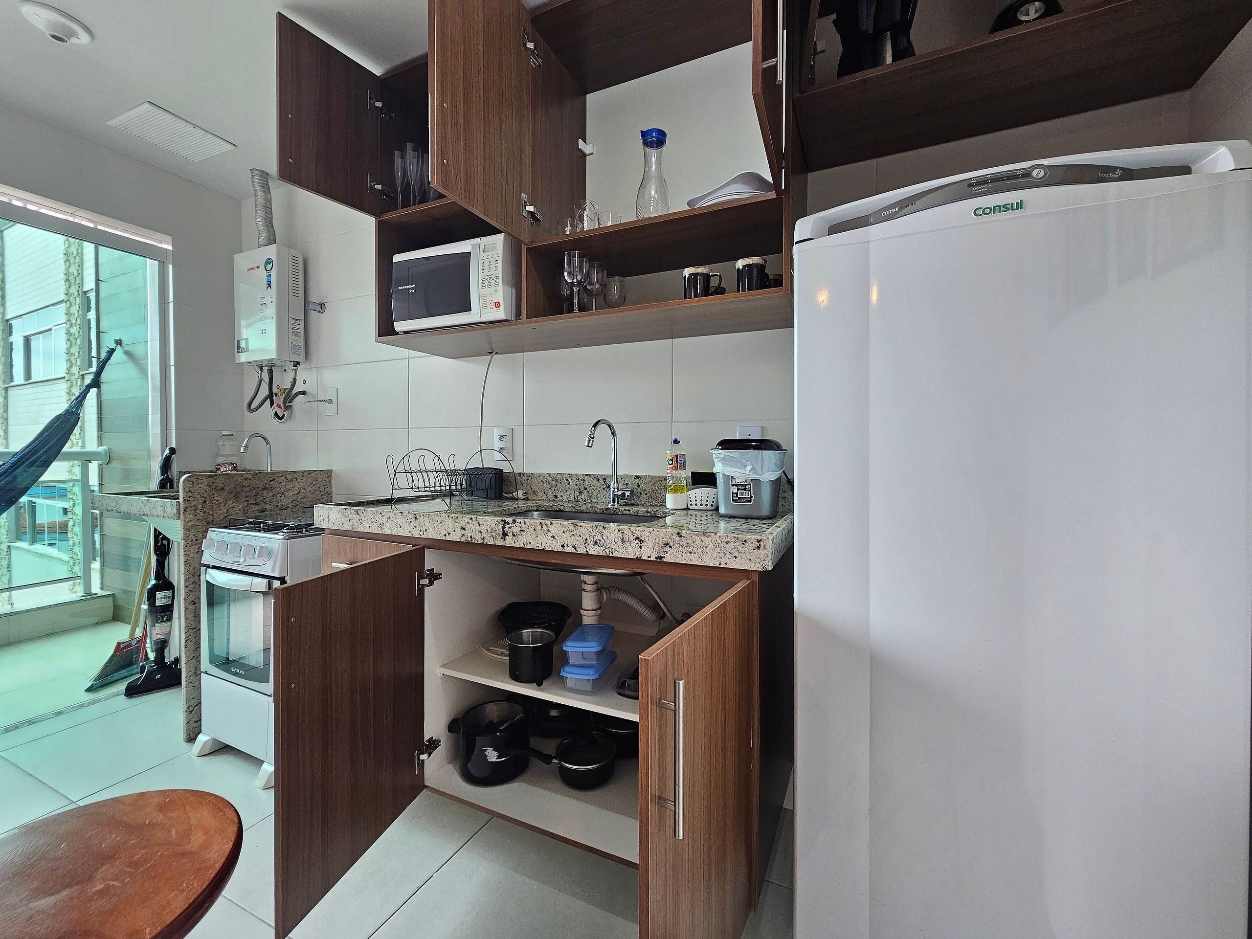 A compact kitchen with a microwave, stove, and refrigerator. There is a window with a view of the outside, and a hammock is visible outside the window.