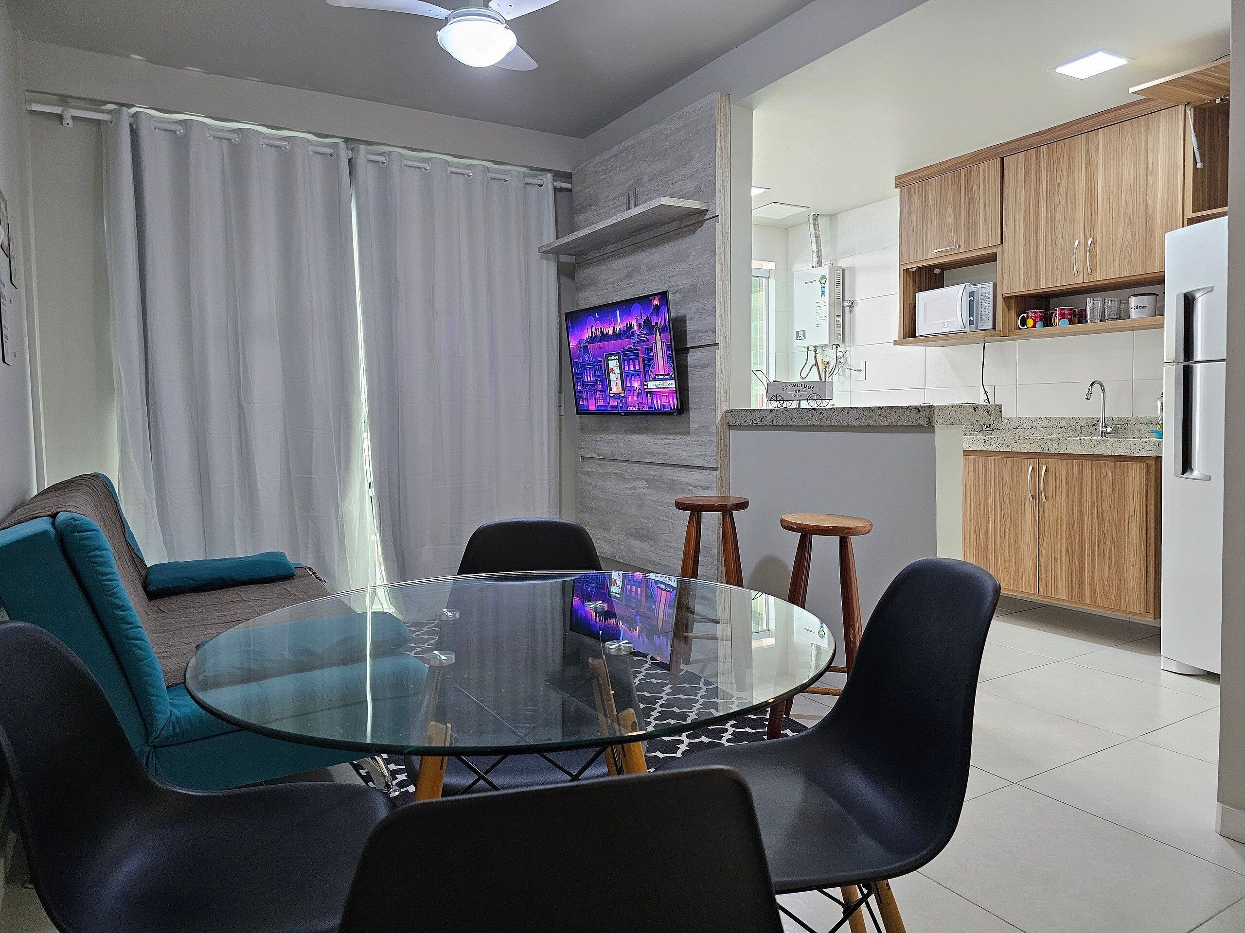 A modern living room with a glass table, black chairs, and a sofa with teal cushions. A flat-screen TV is mounted on the wall.