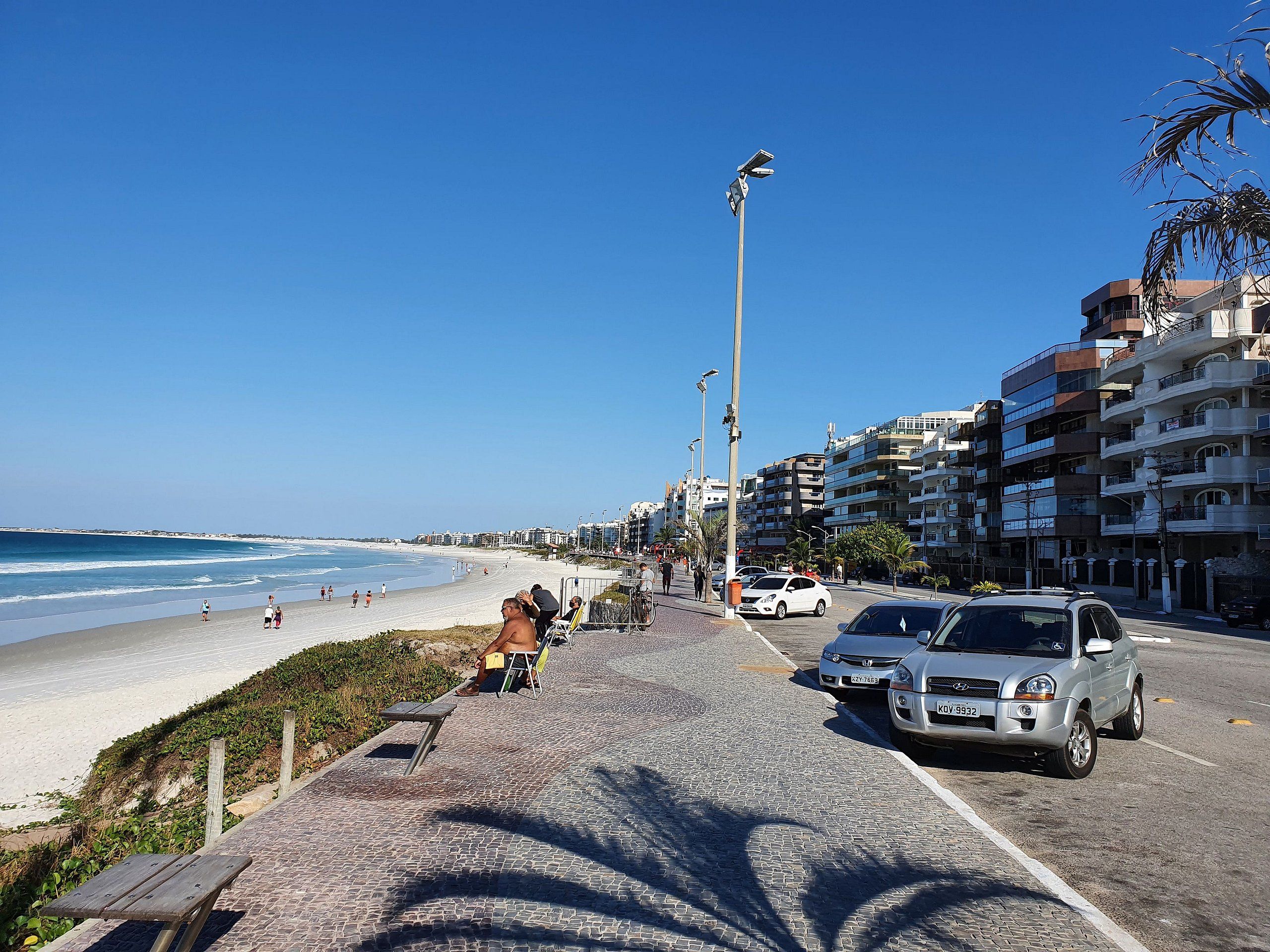 A coastal road with a beach on one side, parked cars, and buildings in the background.