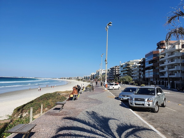 A coastal road with a beach on one side, parked cars, and buildings in the background.