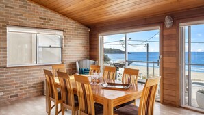 Een eethoek met een houten tafel en stoelen, een bakstenen muur en uitzicht op strand en zee door grote ramen.