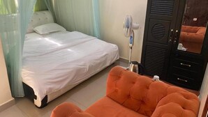 A bedroom with a bed, an orange ottoman, a standing fan, and a wardrobe. - YNM Adam's Hotel (Pallisa)