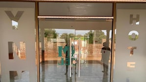 A hotel entrance with glass doors and a visible reflection of a brick wall and greenery outside. - YNM Adam's Hotel (Pallisa)