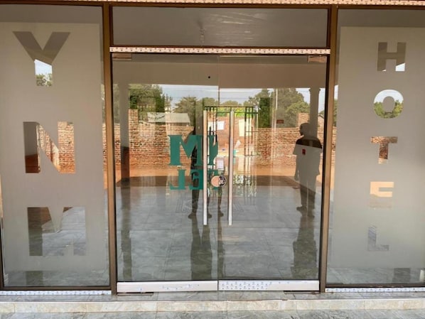Entrada de hotel con puertas de vidrio y el reflejo visible de una pared de ladrillo y follaje en el exterior.
