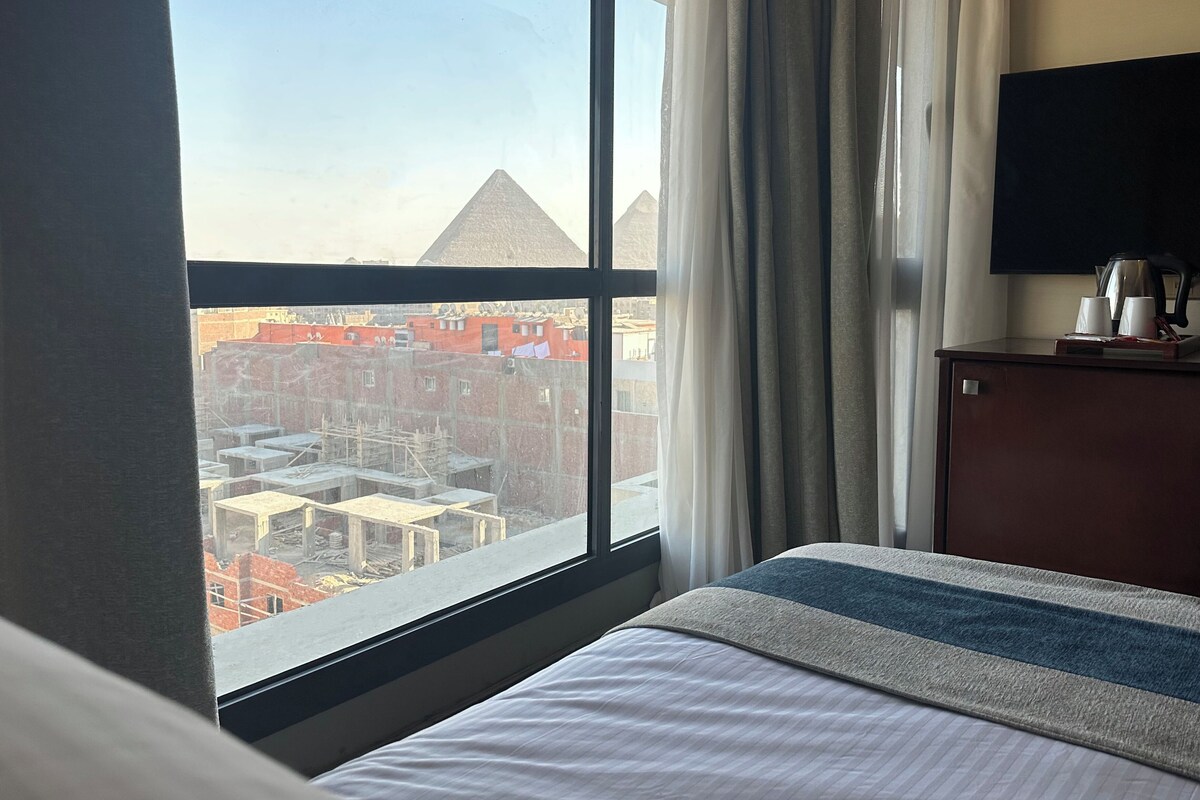 A hotel room with a large window offering a view of pyramids and buildings.