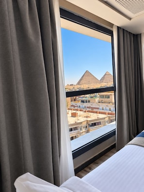 A hotel room with a large window, curtains, a bed, and a view of the pyramids. - Stay Inn Pyramids Hotel (Giza)