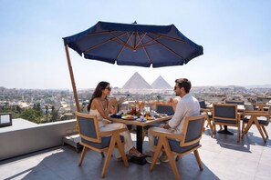 A rooftop dining area with a view of the pyramids, wooden chairs, and a table with food and drinks. - Stay Inn Pyramids Hotel (Giza)