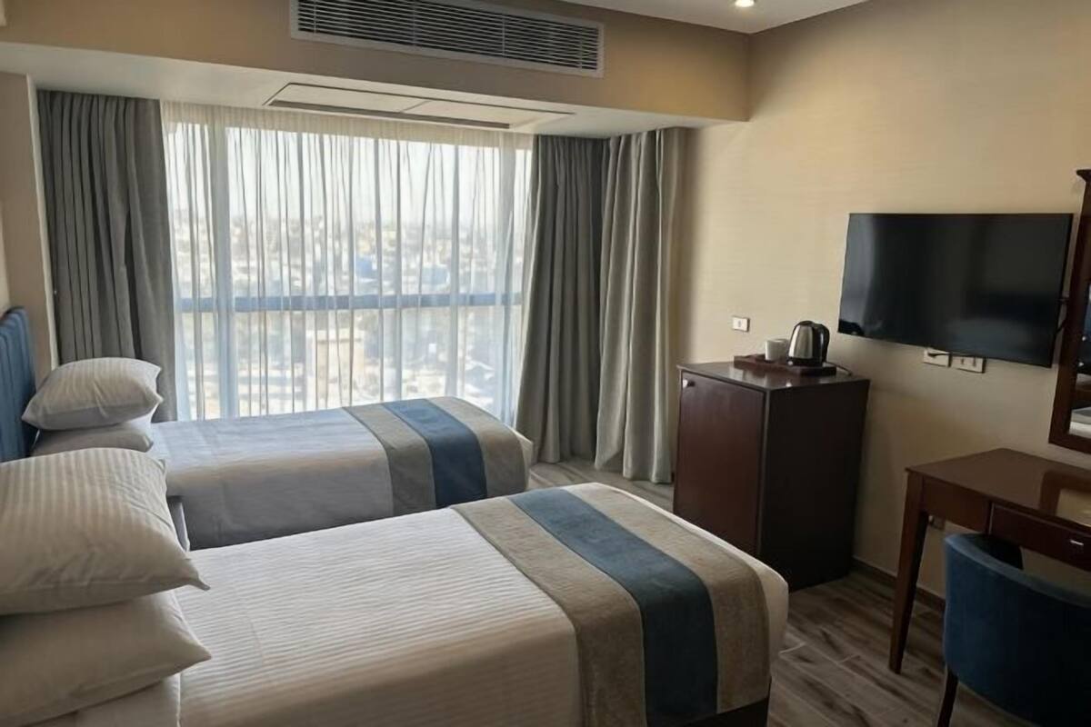 A hotel room with two beds, a flat-screen TV, a desk, and a view of a cityscape through large windows.