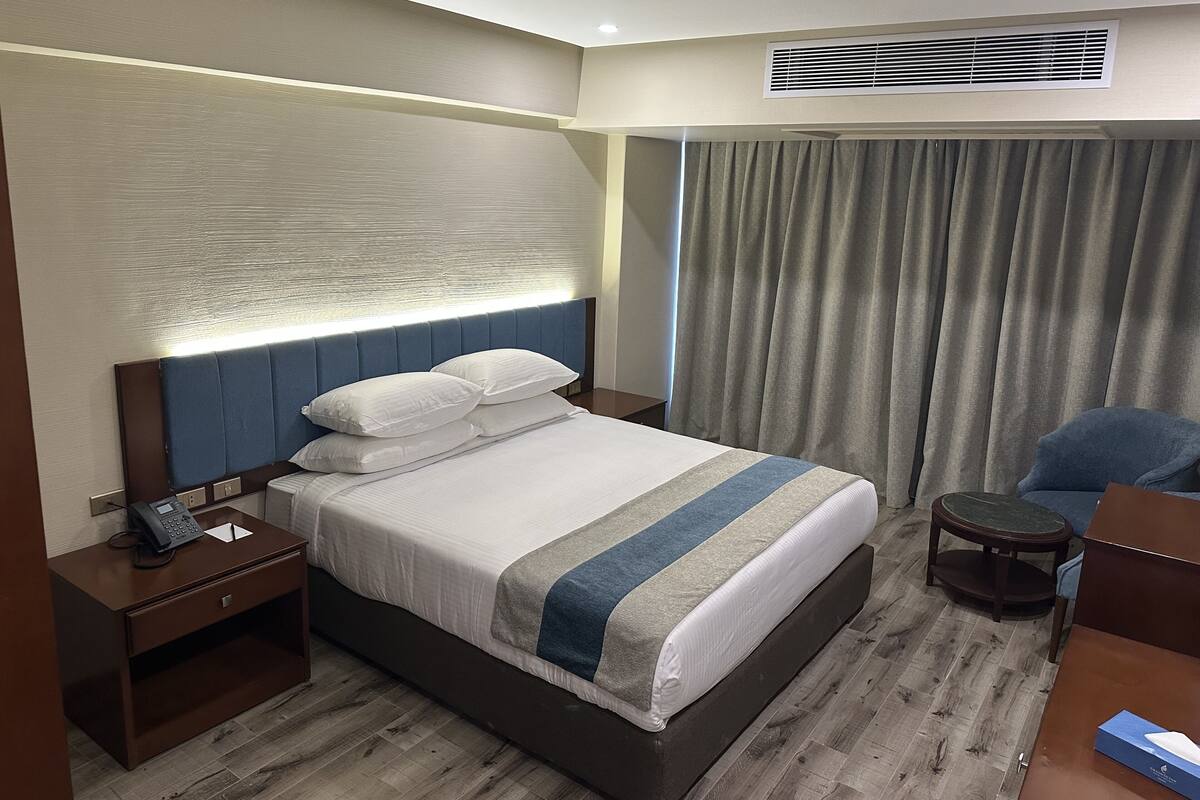 A hotel room with a large bed, a bedside table with a phone, a chair, and a desk.