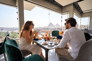 Una pareja cenando en una mesa con vistas a la ciudad, incluyendo pirámides, en un interior moderno con ventanas amplias y sillas verdes.