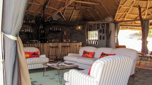 A room with a thatched roof, a bar area with bottles, a white sofa with striped cushions, and a green patterned rug. - Ahadi Mara Camp (Maasai Mara)