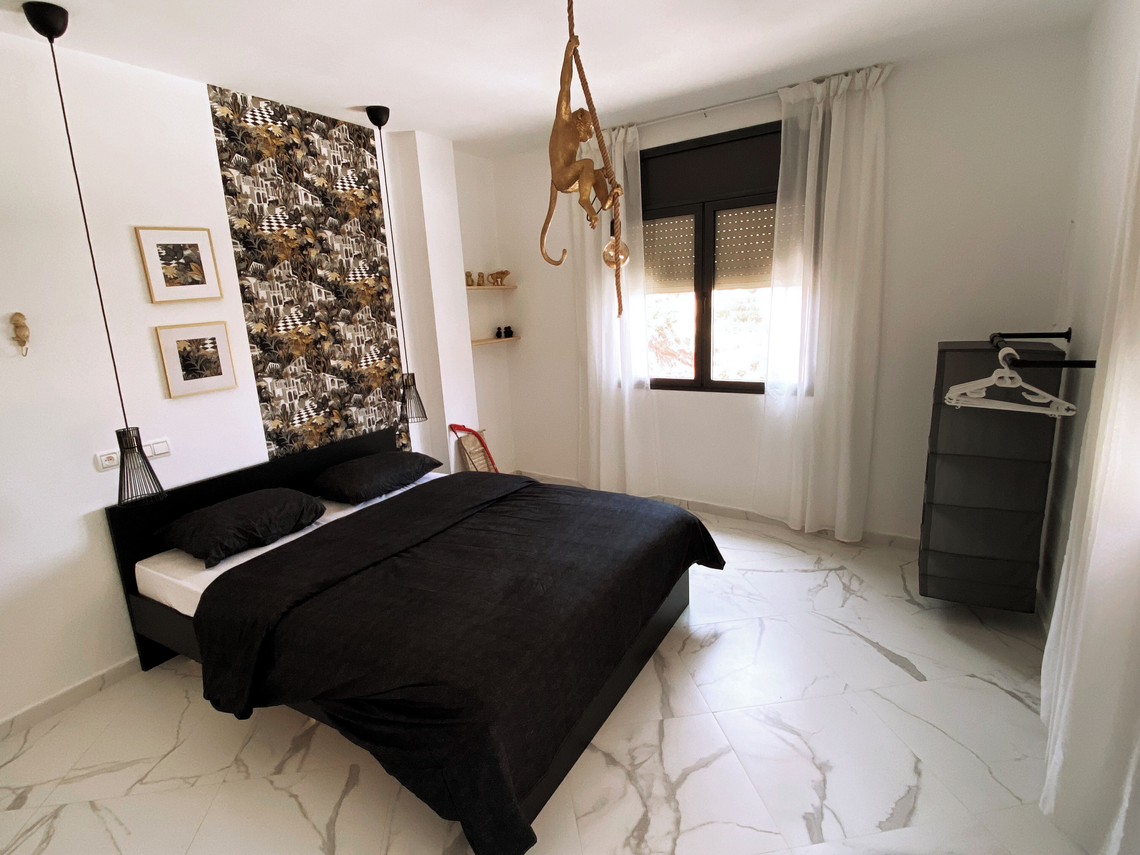 Un dormitorio con cama, papel tapiz estampado en una pared, cortinas en una ventana y una lámpara colgante.