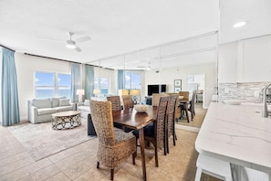 A spacious living room with a large window, a dining area with a wooden table and chairs, and a kitchen with a white countertop.