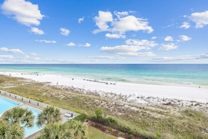 A beach view with a clear sky, a sandy beach, and a pool area with lounge chairs.