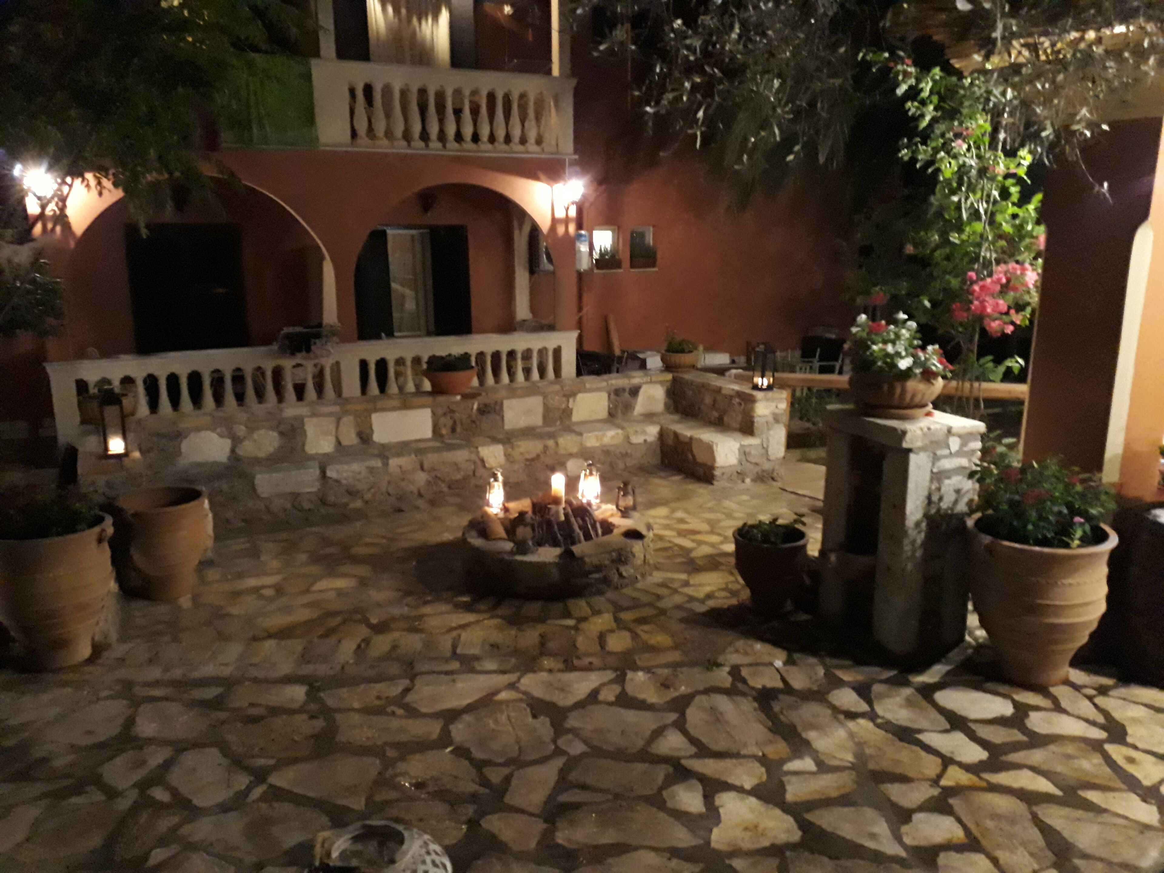 An outdoor courtyard with stone flooring, a central fire pit, potted plants, and a stone wall with a balcony.