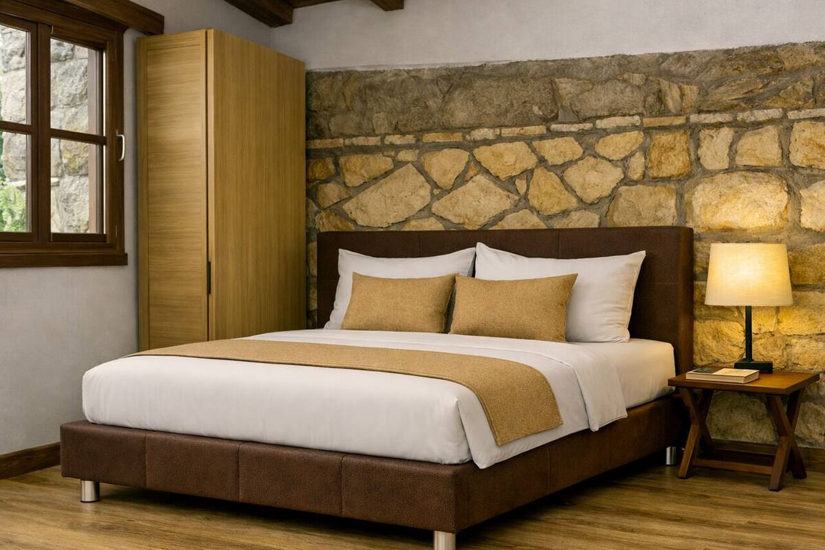 A bedroom with a stone wall, a wooden wardrobe, a bed with a brown headboard, and a bedside table with a lamp.