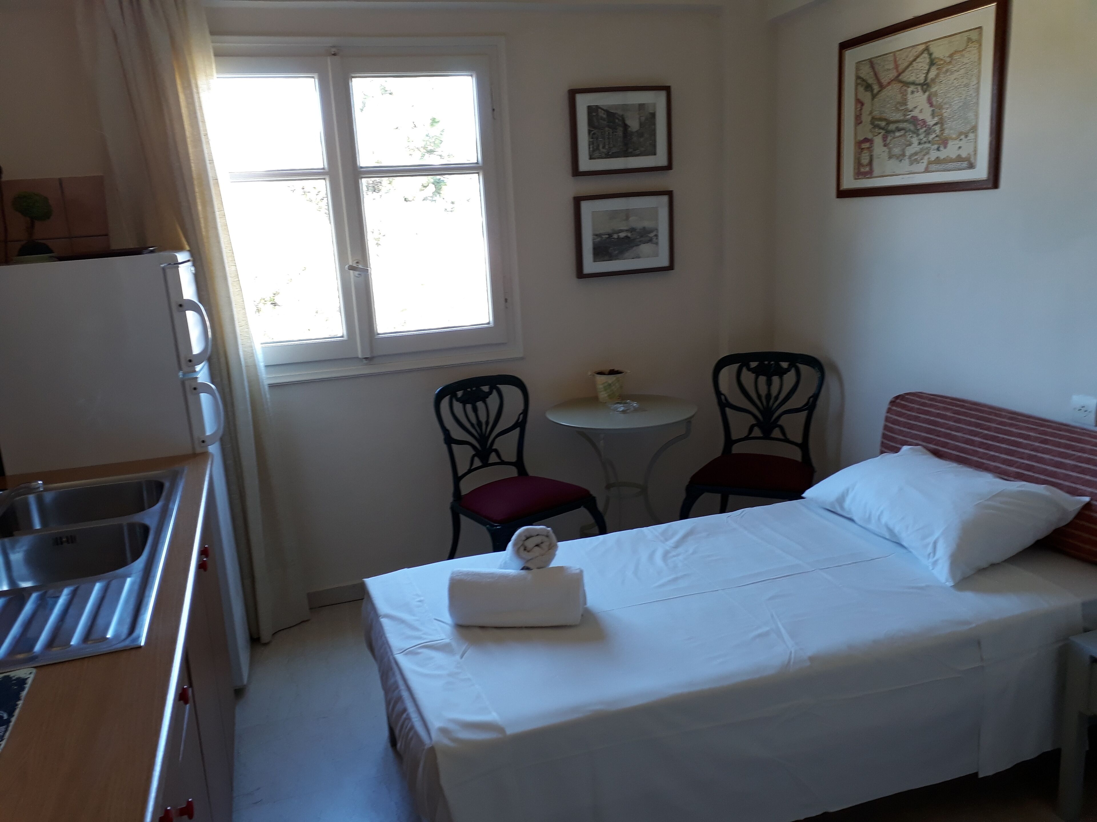 Una habitación pequeña y bien iluminada con una cama, una mesa con sillas, un ventanal y un rincón de cocina.