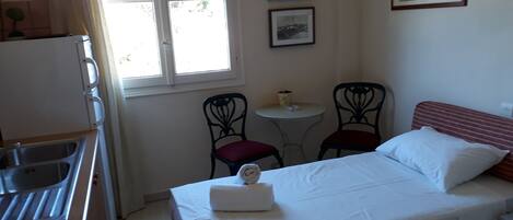 Una habitación pequeña y bien iluminada con una cama, una mesa con sillas, un ventanal y un rincón de cocina.