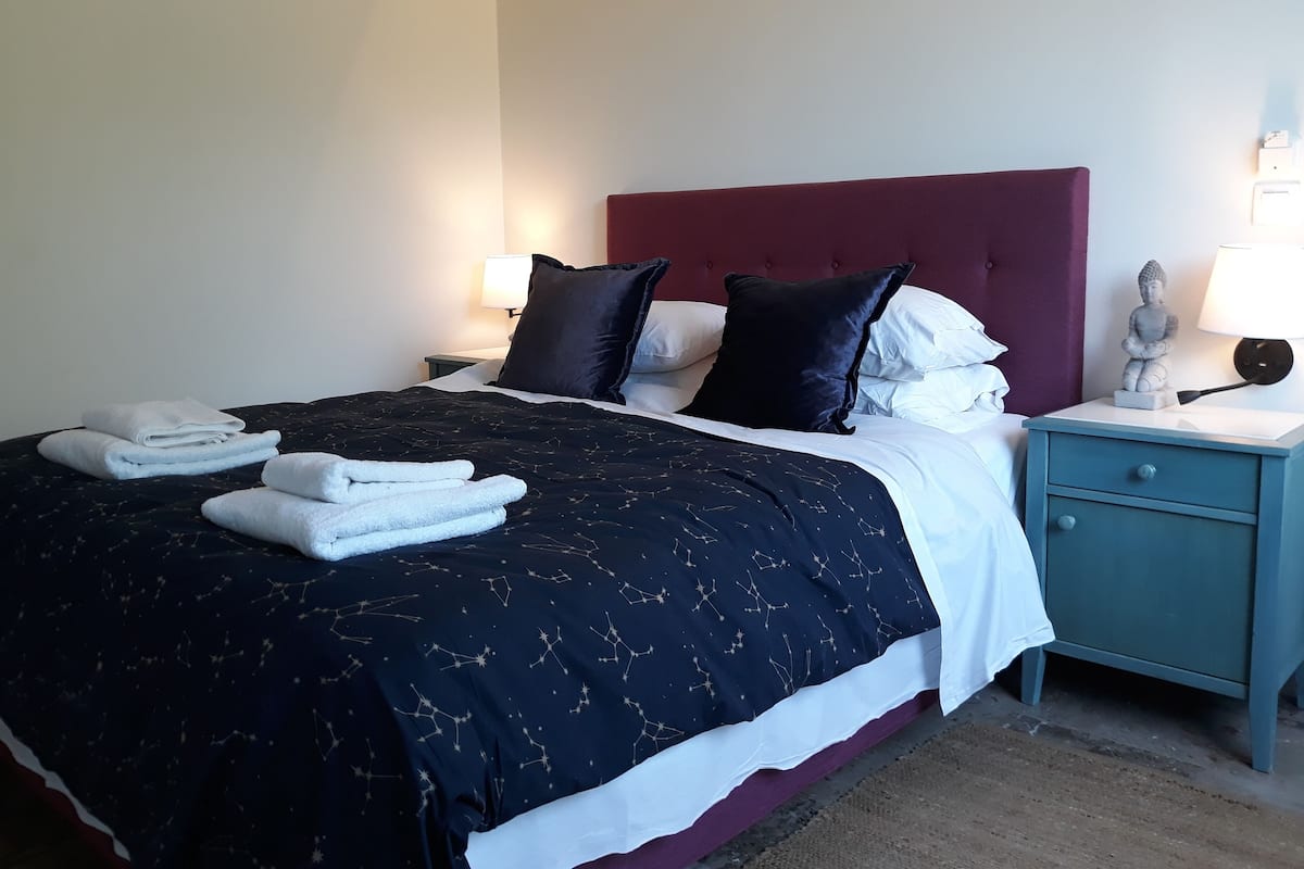 A neatly made bed with a dark patterned bedspread, white pillows, and folded towels on the side. A small nightstand with a lamp and a statue is beside the bed.
