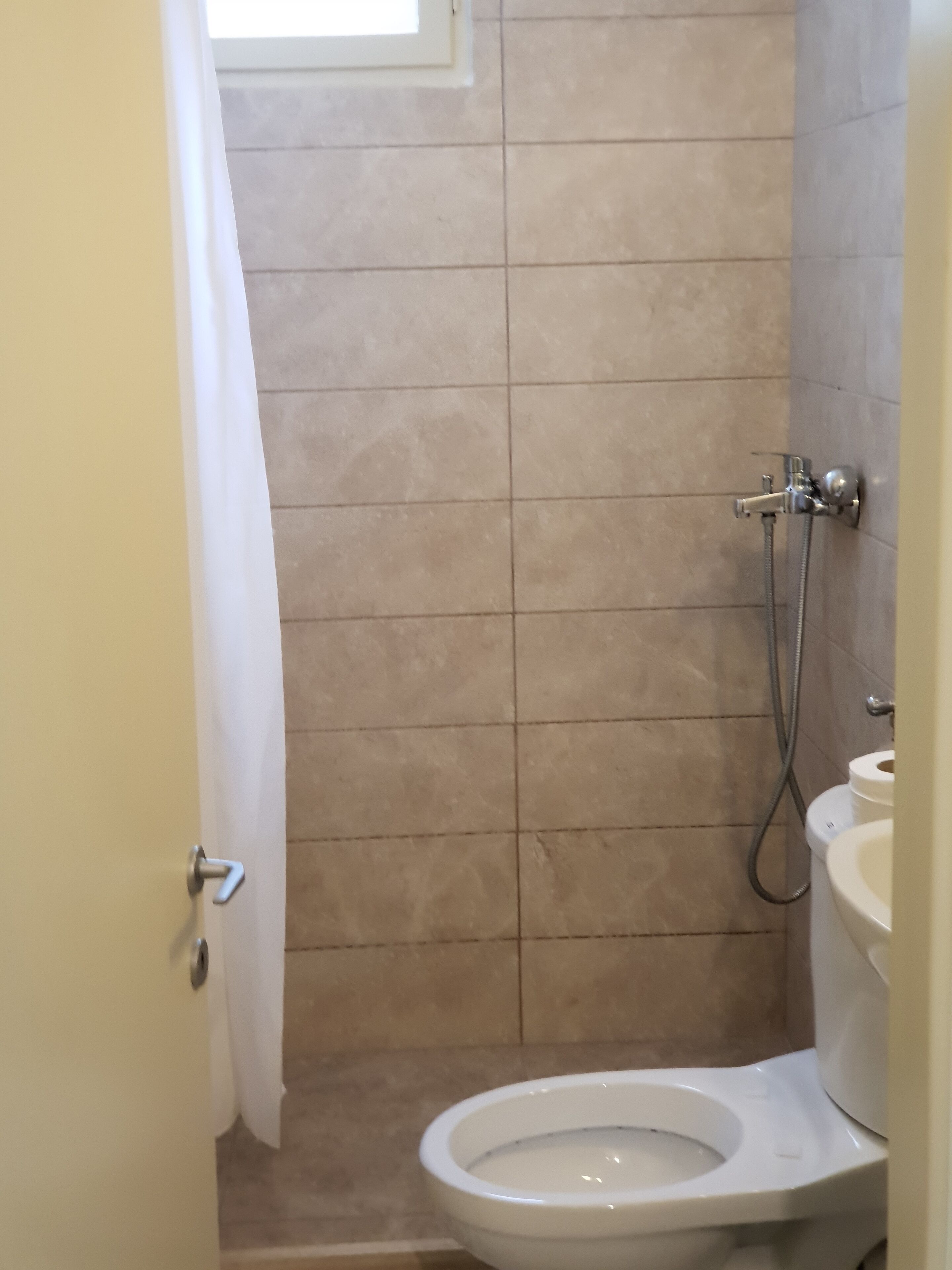 A small bathroom with a toilet, a shower, and a window with a curtain.