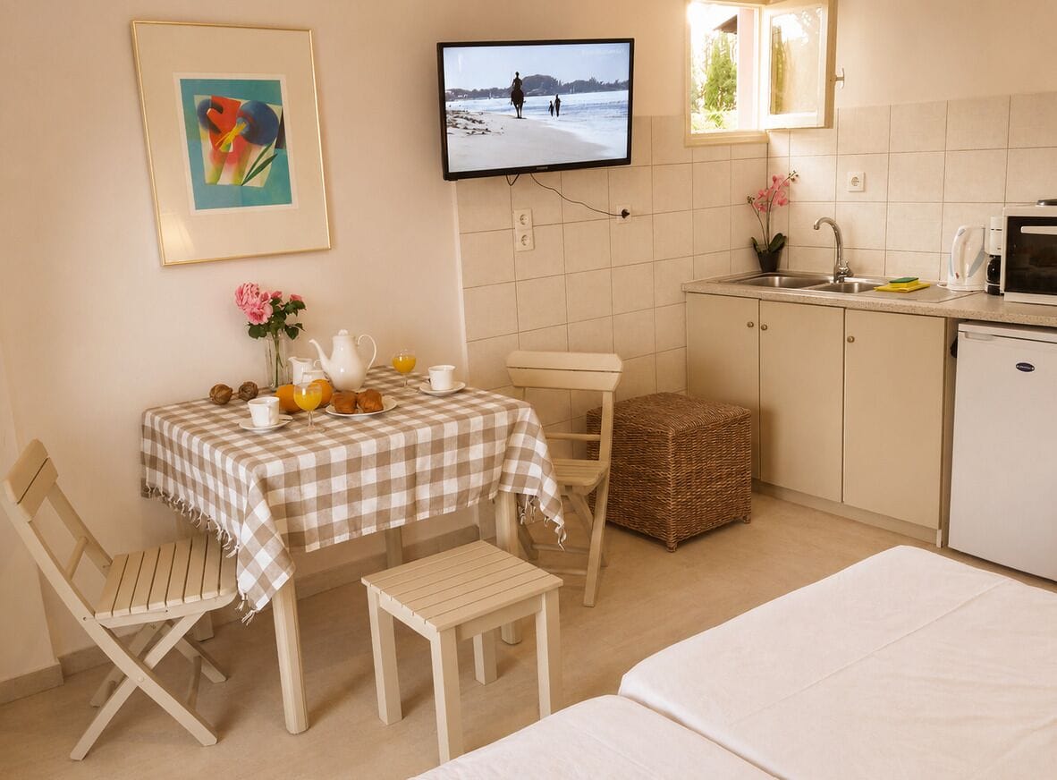 Una habitación pequeña y limpia con una cama, una mesa con cosas para el desayuno, un televisor mostrando una escena de playa, una cocina y una silla de mimbre.