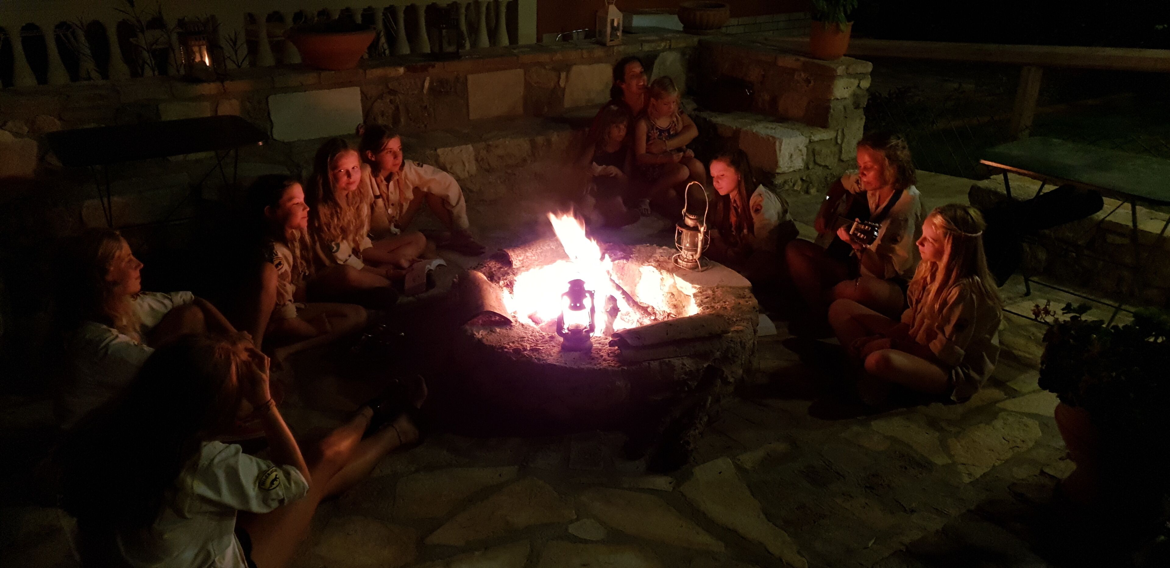 Un grupo de personas reunidas alrededor de una fogata en un entorno exterior con paredes de piedra y macetas con plantas.