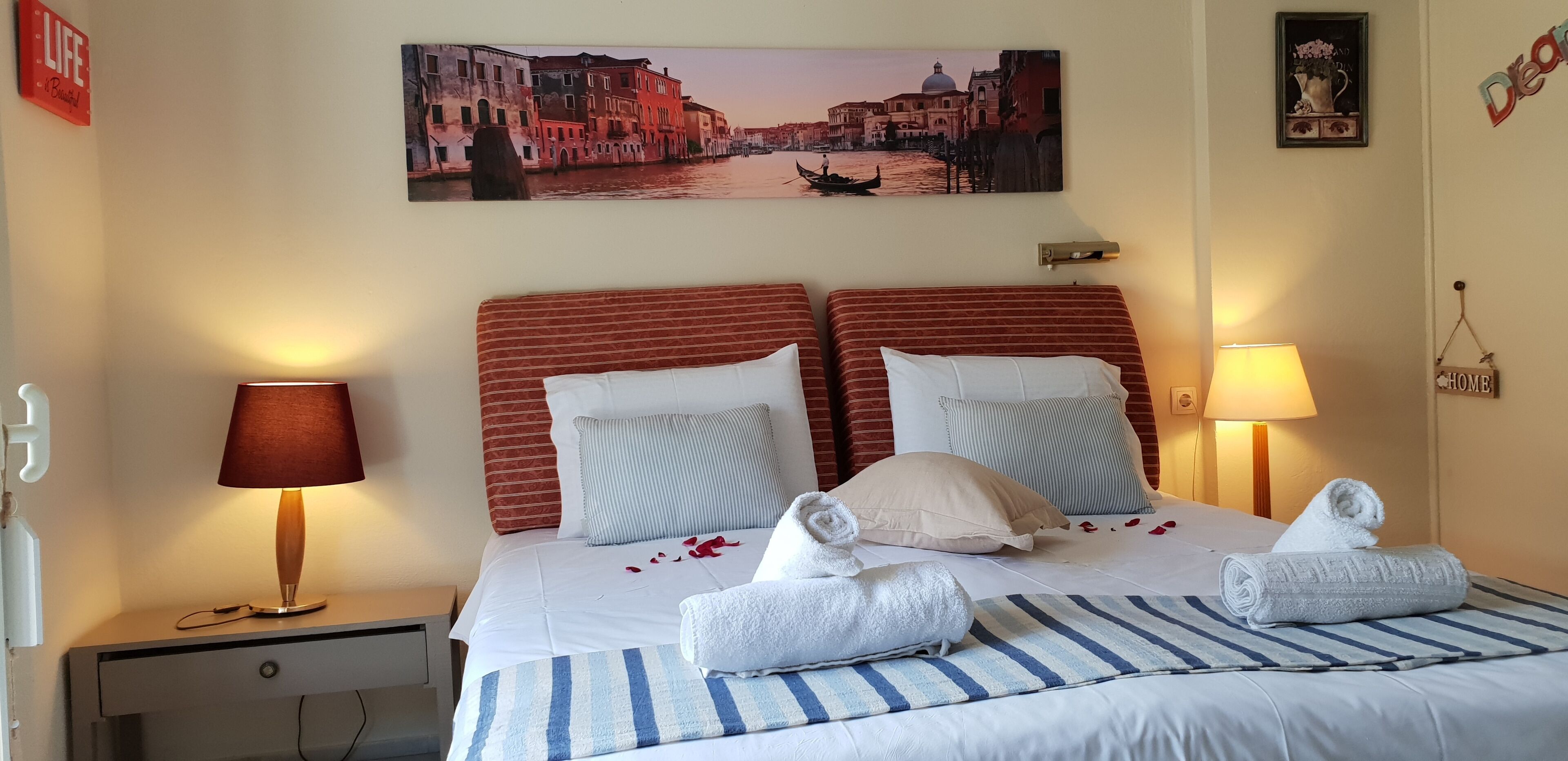 Quarto de hotel com cama, roupa de cama listrada, duas almofadas e arranjo decorativo de toalhas. Uma pintura retratando uma cena de canal está acima da cama, e há duas luminárias de cabeceira.