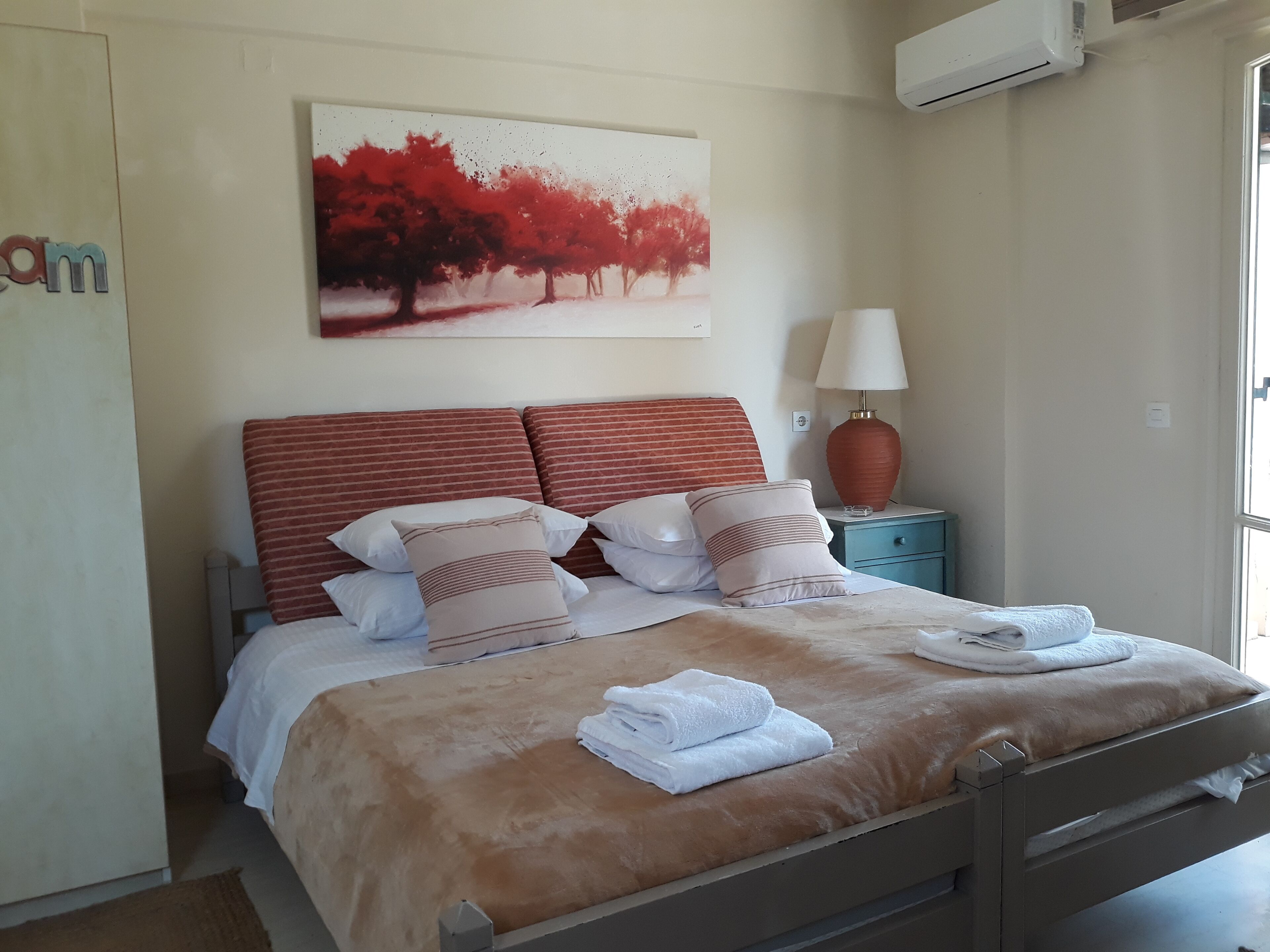 A neatly made bed with a brown cover, white pillows, and folded white towels. There is a painting of red trees on the wall above the bed.