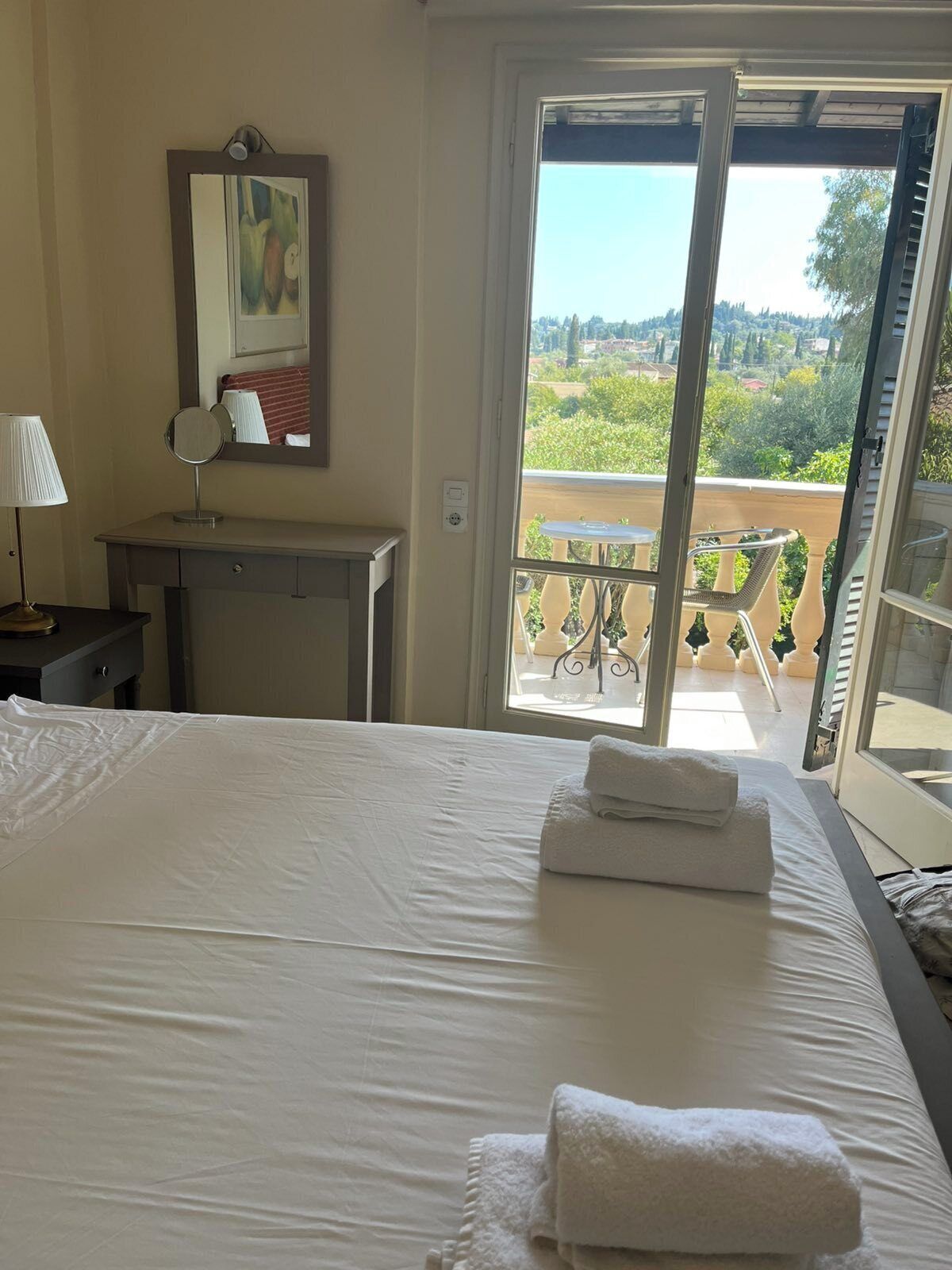 A hotel room with a bed, a desk with a lamp, a mirror, and a balcony with a table and chairs.