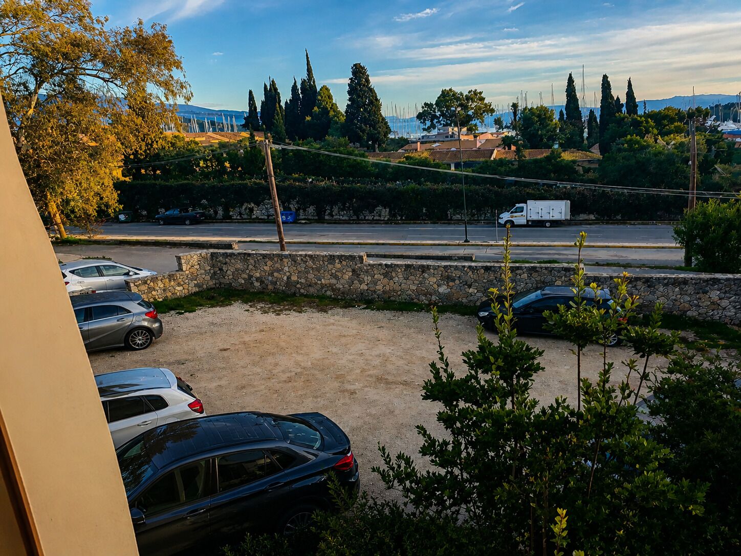 A parking area with several cars, a stone wall, and a view of a road with vehicles and trees in the background.