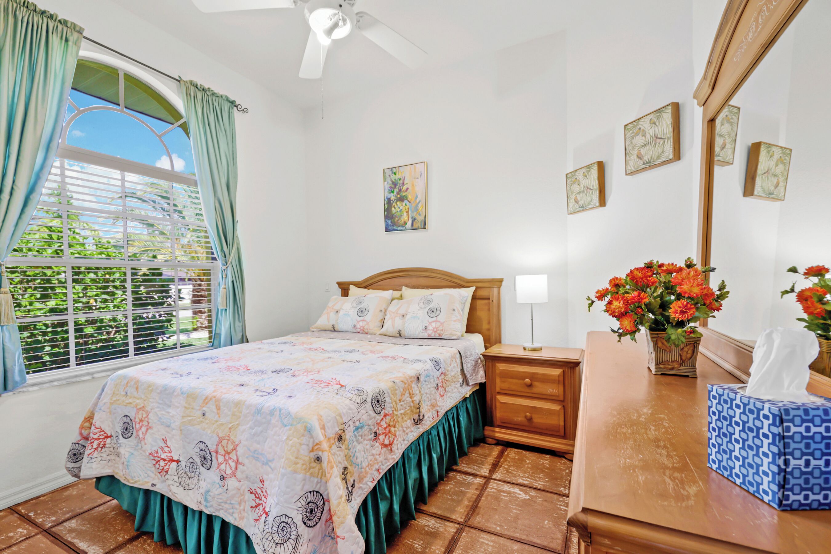 Une chambre à coucher comprenant un lit, une table de chevet, une commode, une fenêtre avec des rideaux et un ventilateur de plafond.