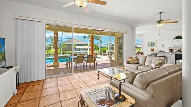 A spacious living room with a large glass door leading to an outdoor pool area, a ceiling fan, and a modern kitchen in the background. - Cape Coral Waterfront Vacation Rental with Heated Pool and Boat Dock. (Cape Coral)