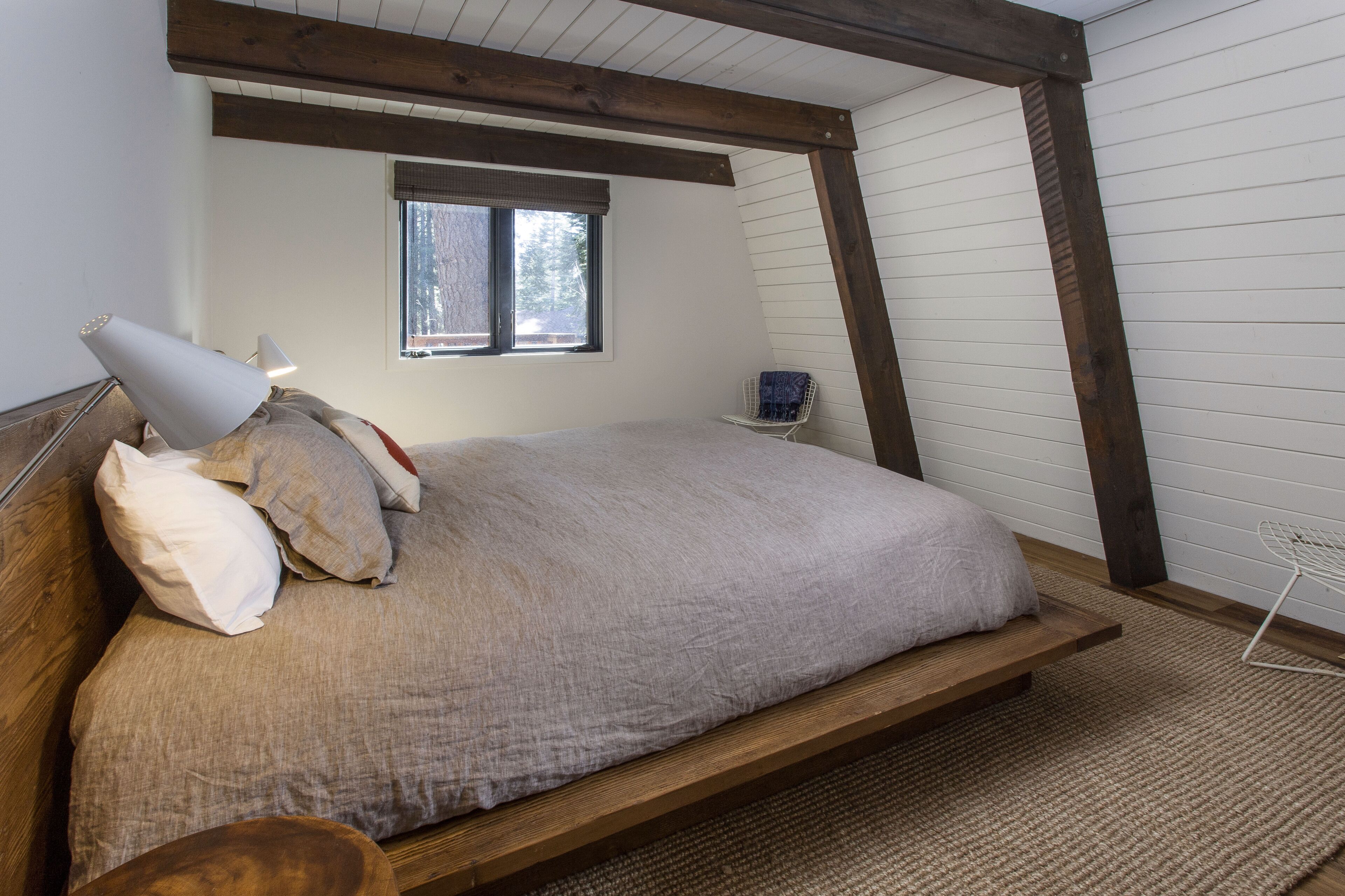 Quarto com cama de madeira, janela com persianas, uma cadeira e uma mesa pequena.