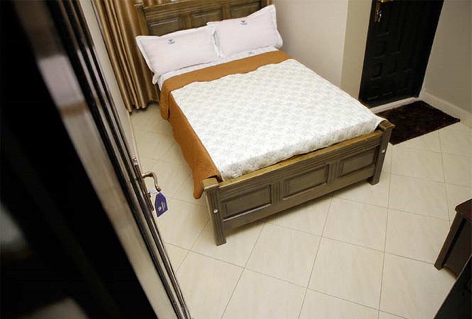 A bed with a white quilt and brown bedspread in a room with a tiled floor and a wooden door.
