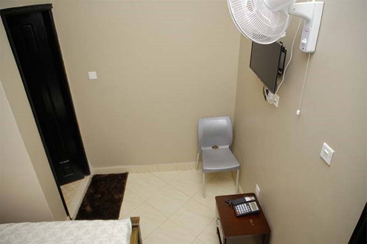 A small room with a bed, a chair, a desk with a keyboard, a fan, and a TV mounted on the wall.