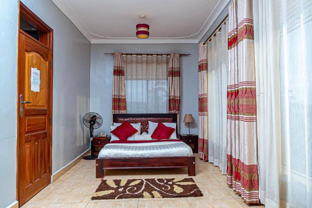 A bedroom with a wooden bed, red and white curtains, a standing fan, and a patterned rug.