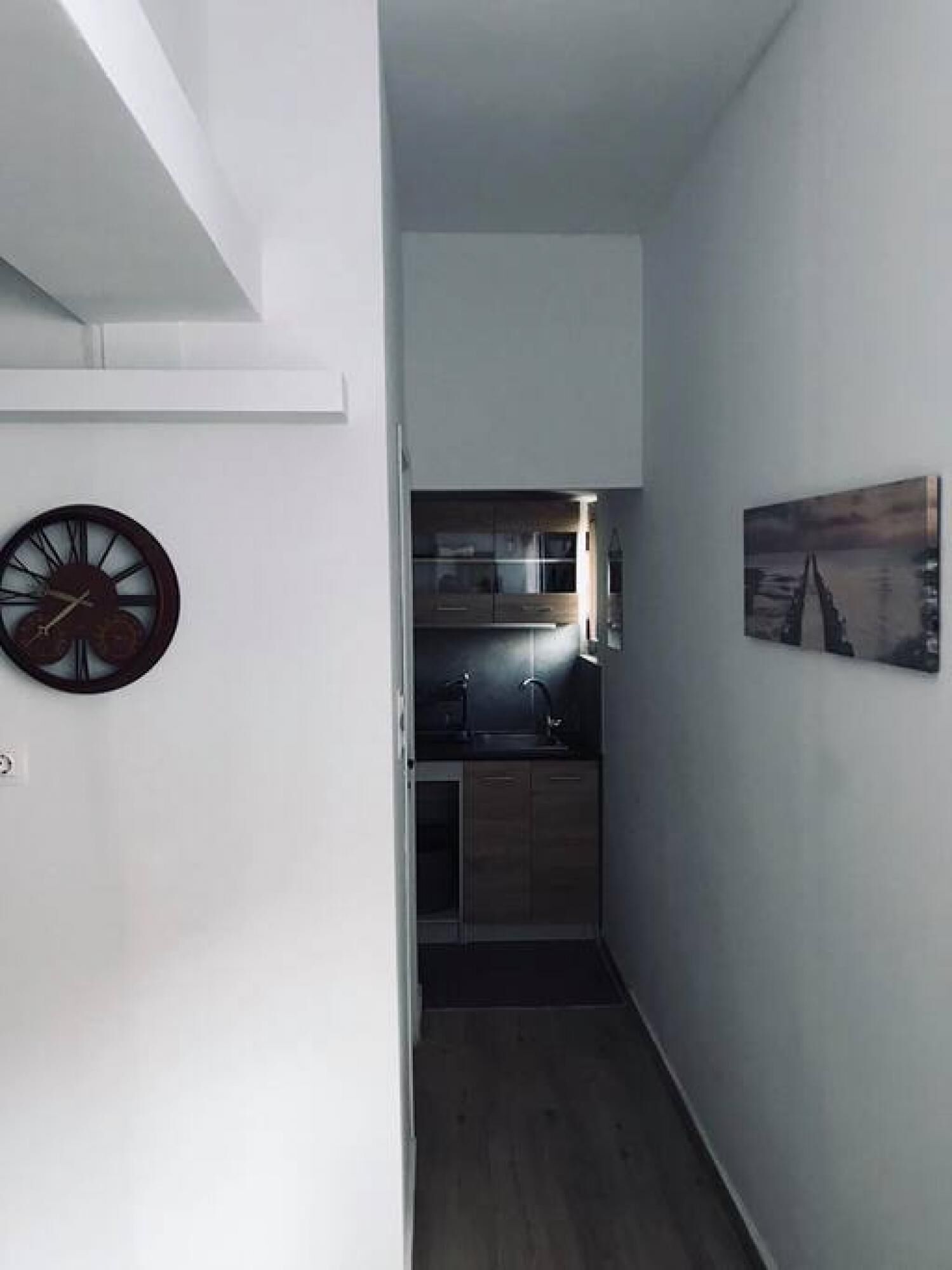 A narrow hallway leading to a kitchen with a clock and a painting on the wall.