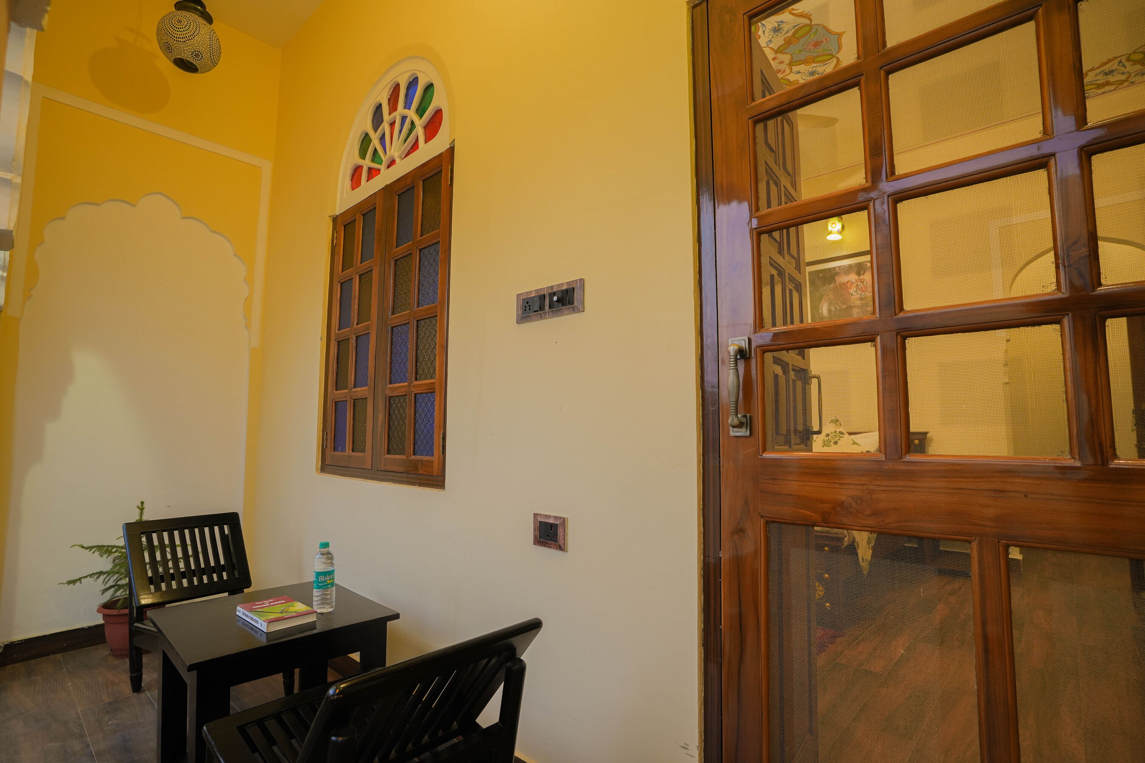 A room with a wooden door, a window with a decorative design, a table with a chair, and a potted plant.