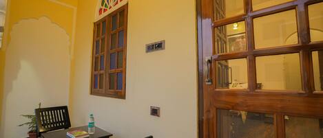 A room with a wooden door, a window with a decorative design, a table with a chair, and a potted plant.