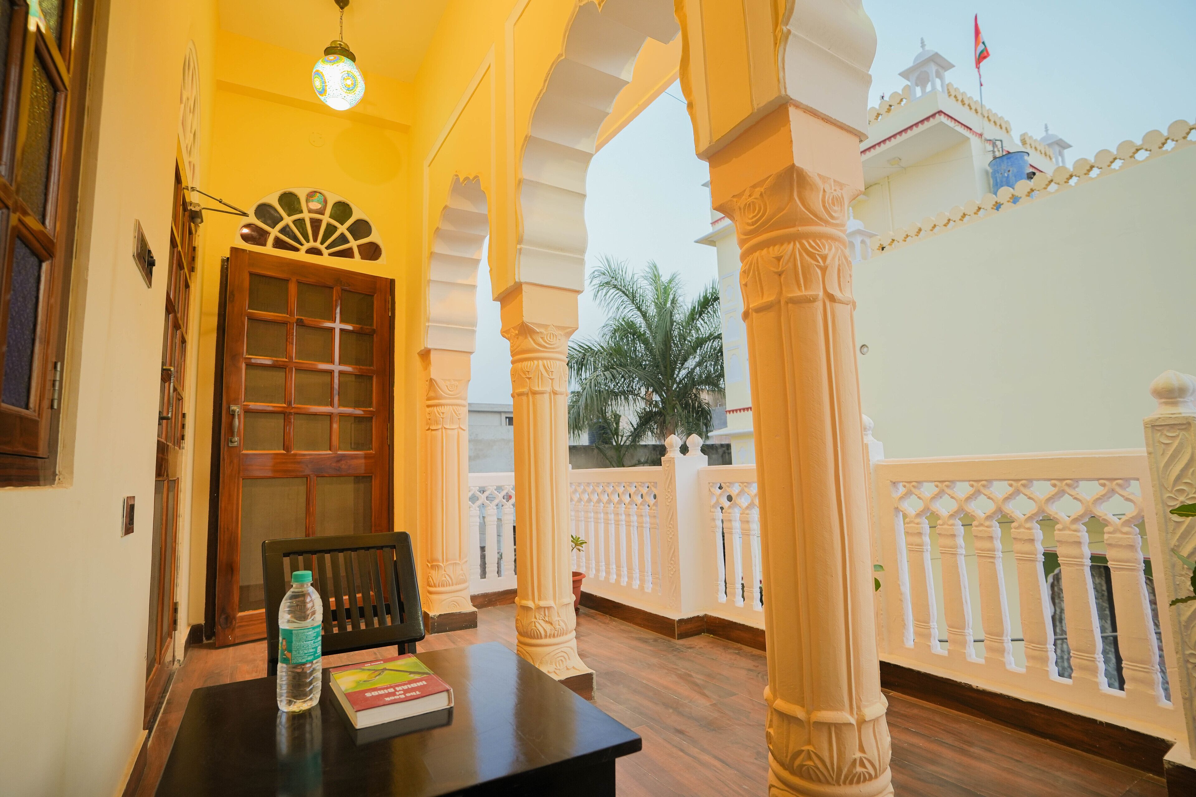 A corridor with a wooden door, a table with a water bottle and a book, and a balcony with white railings and palm trees.