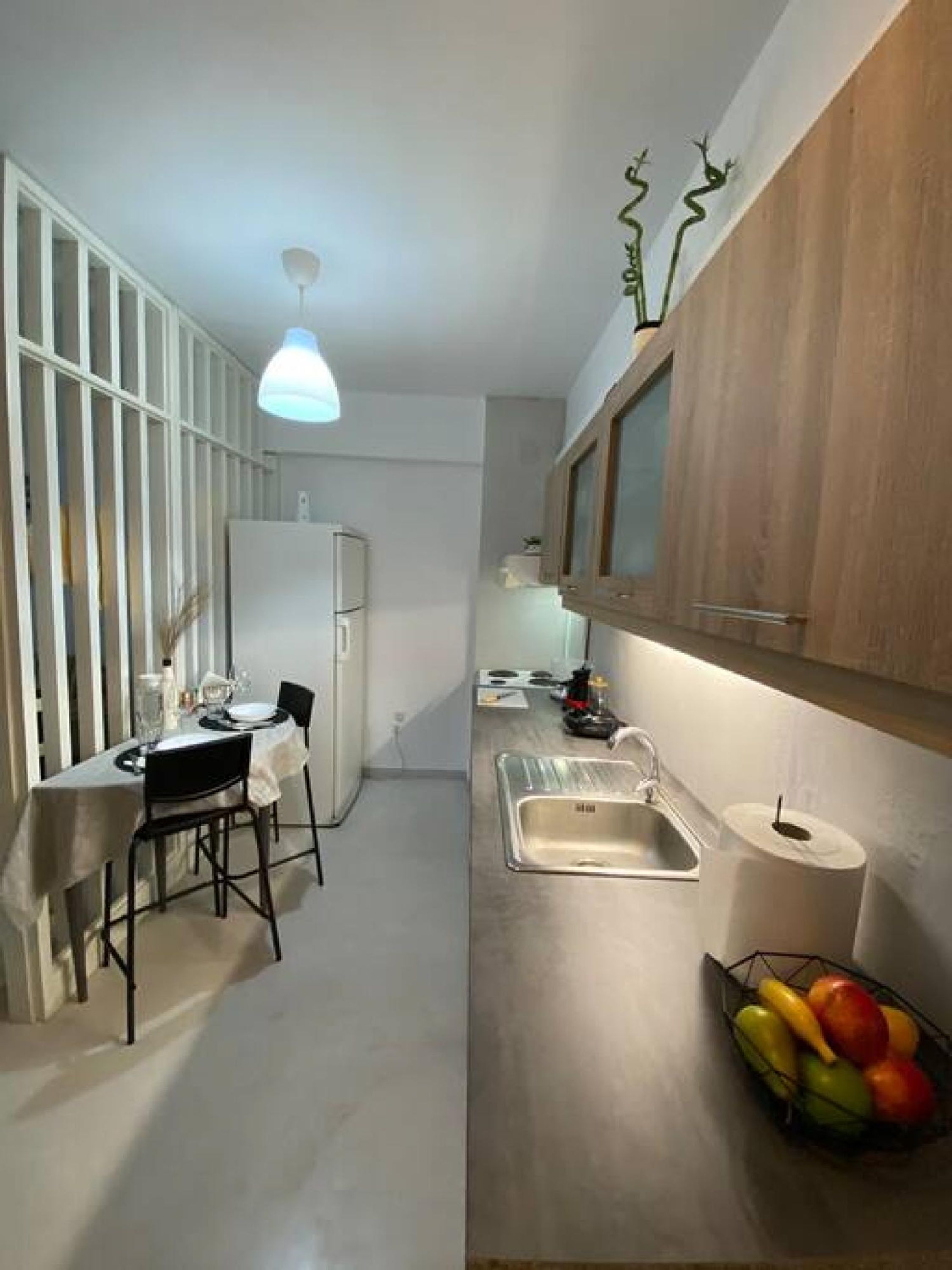 A compact kitchen with a dining area, a built-in refrigerator, a wooden cabinet, a stainless steel sink, and a fruit basket.