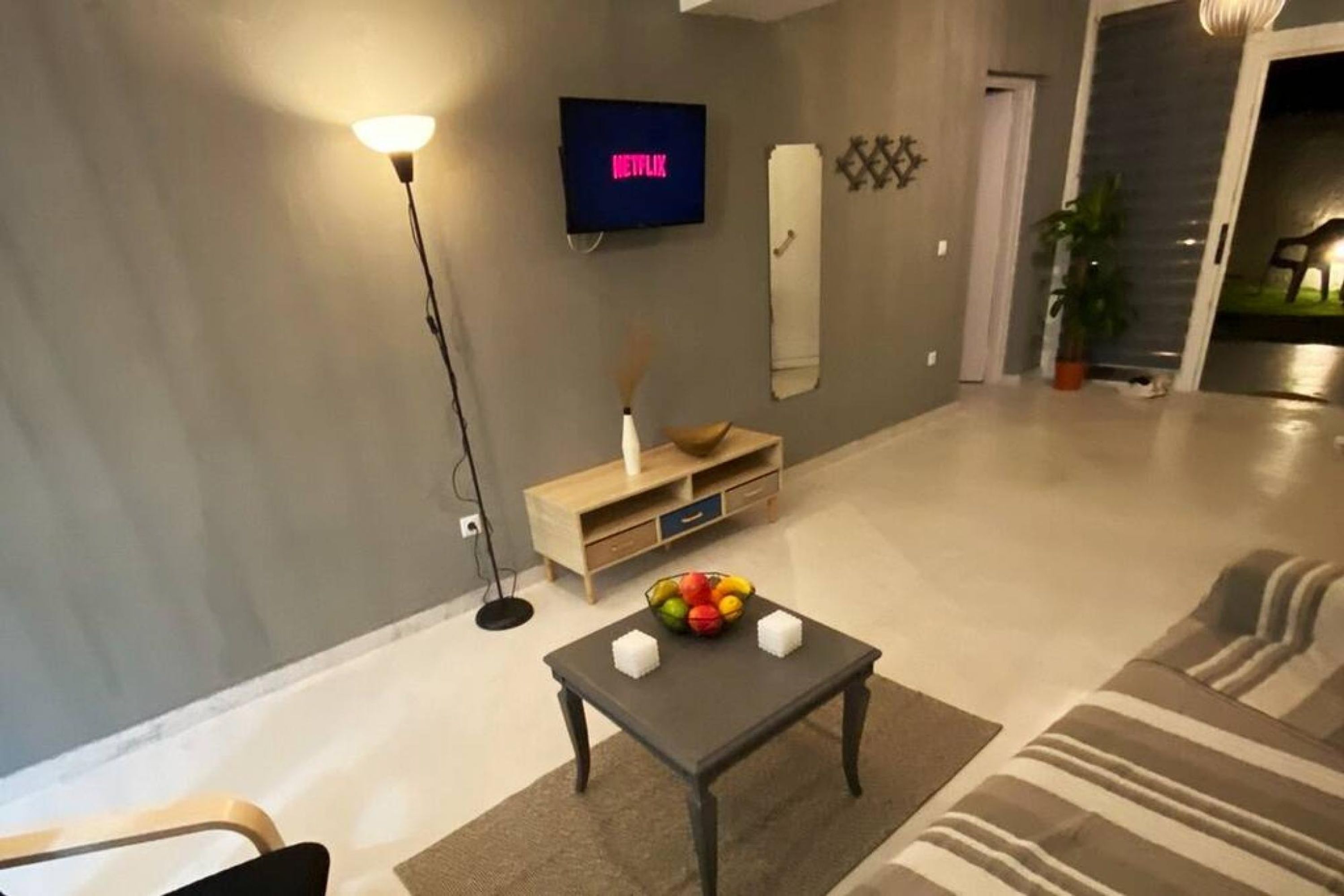 A modern living room with a flat-screen TV mounted on the wall, a small wooden side table with a bowl of fruit, and a striped rug on the floor.