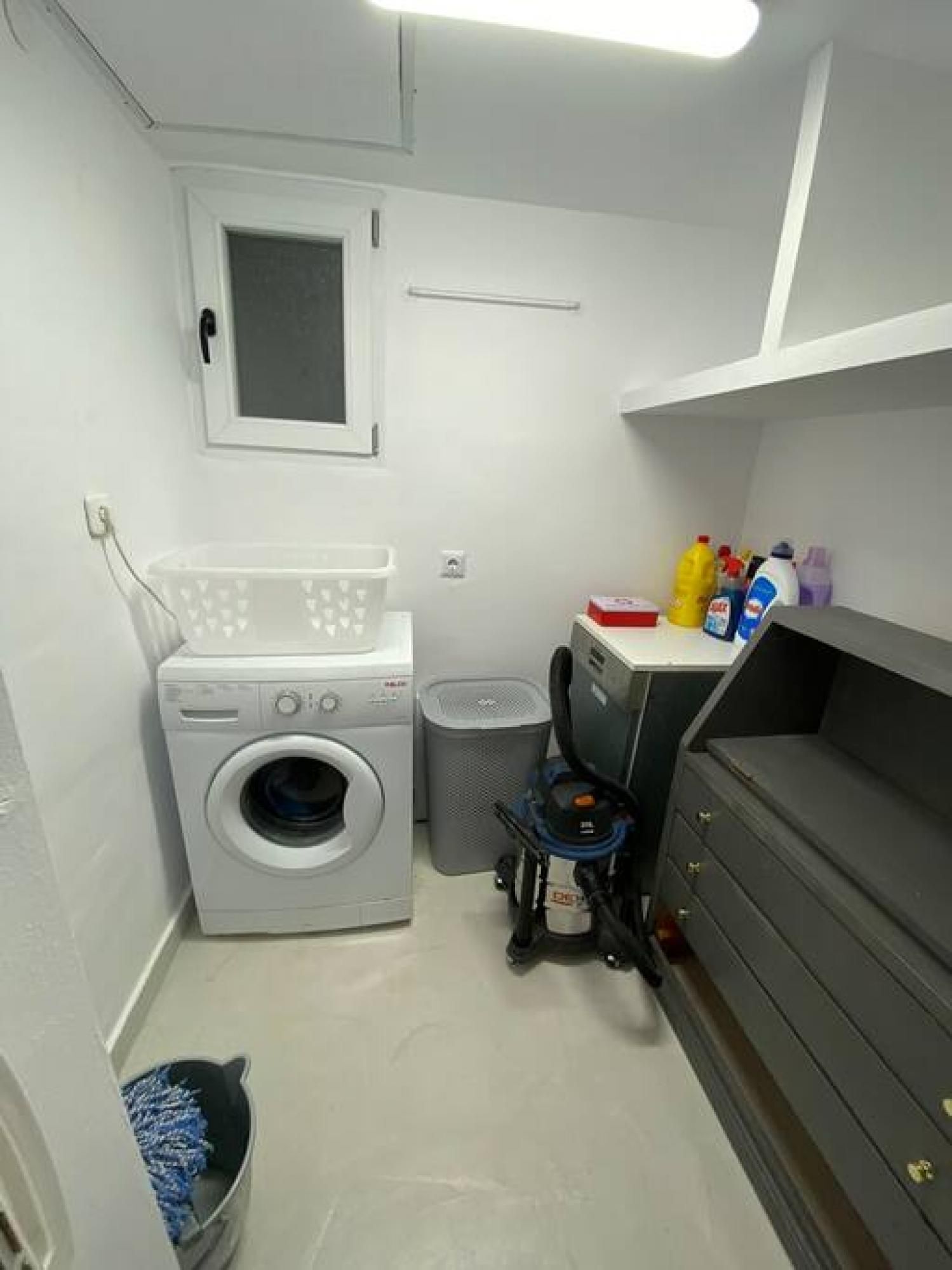 A laundry room with a washing machine, a dryer, a trash bin, and a shelf with cleaning supplies.