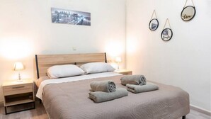 Un lit bien fait, recouvert d’une courtepointe beige, avec deux lampes de chevet et un tableau représentant un paysage urbain accroché au mur.