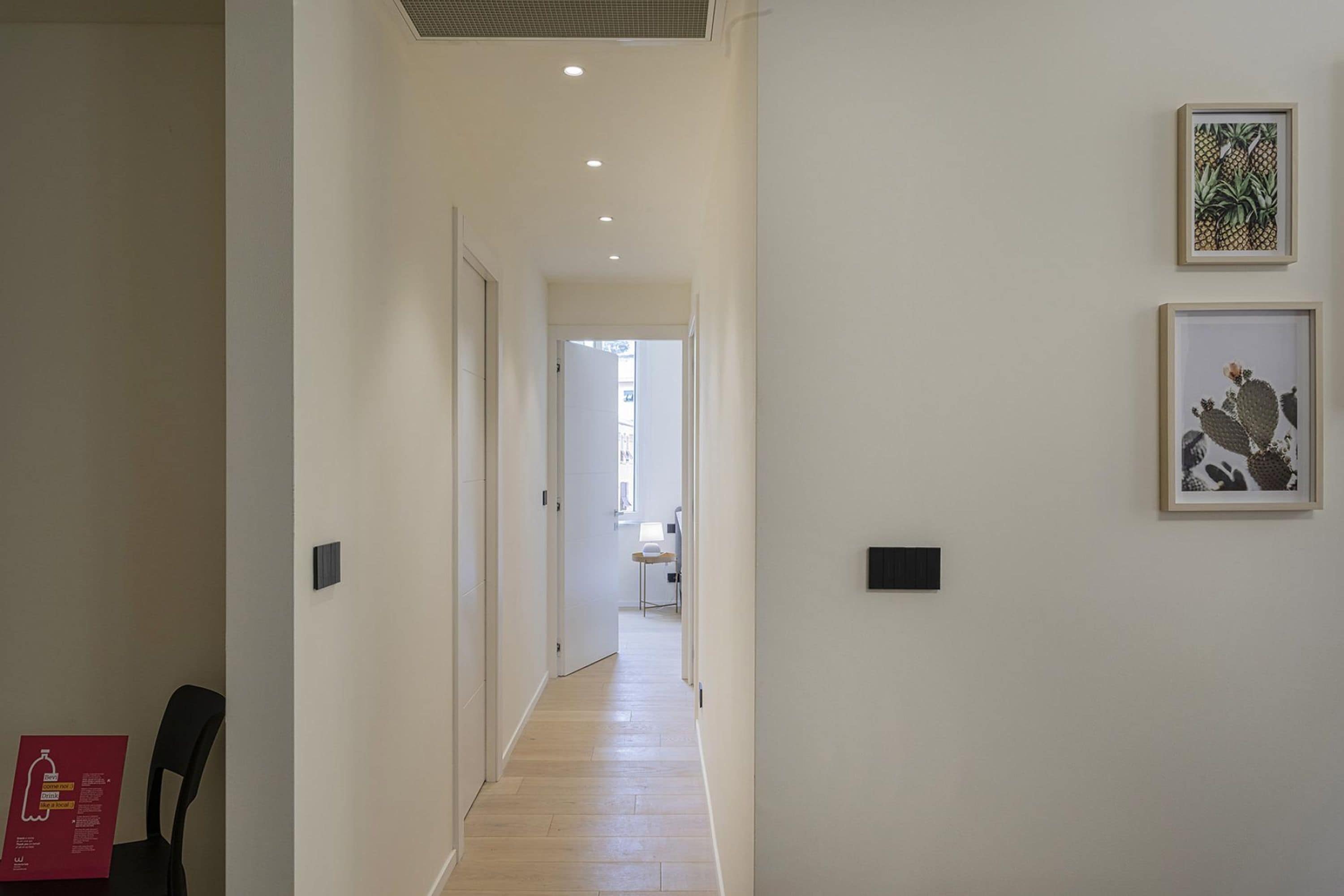 Un pasillo con piso de madera, paredes blancas y dos cuadros enmarcados. Hay una puerta al final del pasillo.