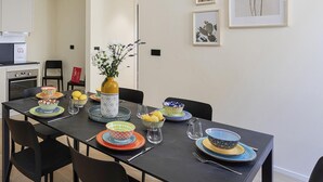 A dining area with a dark table set for four, surrounded by chairs. There are framed botanical prints on the wall, a vase with flowers on the table, and a kitchen area visible in the background. - Riviera Flavour Apartments by Wonderful Italy - Alloro (Santa Margherita Ligure)