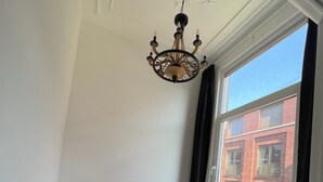 Una habitación con techos altos, un candelabro, un ventanal con vistas a un edificio de ladrillo rojo, y un banco con cojín blanco.