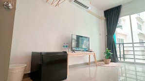 A modern hotel room with a flat-screen TV on a wooden stand, a small refrigerator, a trash bin, and a potted plant near a large window with a view of buildings. - BAOBAB (Patong)