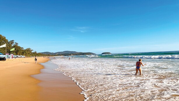 A beach with a person standing in the water, a sandy shore, and distant hills. - BAOBAB (Patong)