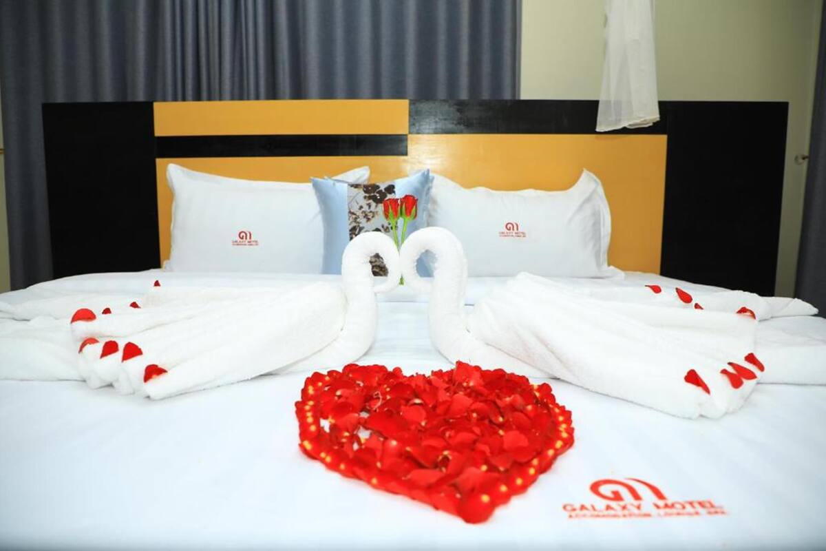 Habitación de hotel con cama, toallas blancas, pétalos de rosa y un logotipo en la cabecera.